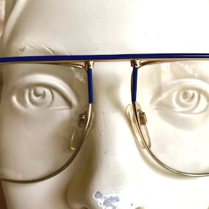 May include: Gold-rimmed eyeglasses with blue accents on the top bar and temples. The glasses have a classic aviator shape with clear lenses and nose pads. The frame is set against a white background.
