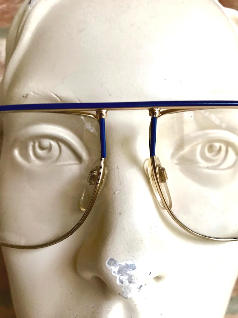 May include: Gold-rimmed eyeglasses with blue accents on the top bar and temples. The glasses have a classic aviator shape with clear lenses and nose pads. The frame is set against a white background.