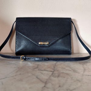 Könnte beinhalten: Eine schwarze Umhängetasche mit einem strukturierten, reptilienartigen Muster. Die Tasche hat einen Klappenverschluss mit einem goldfarbenen Verschluss und einen langen, verstellbaren Riemen. Die Tasche ist rechteckig und hat ein klassisches, elegantes Design.