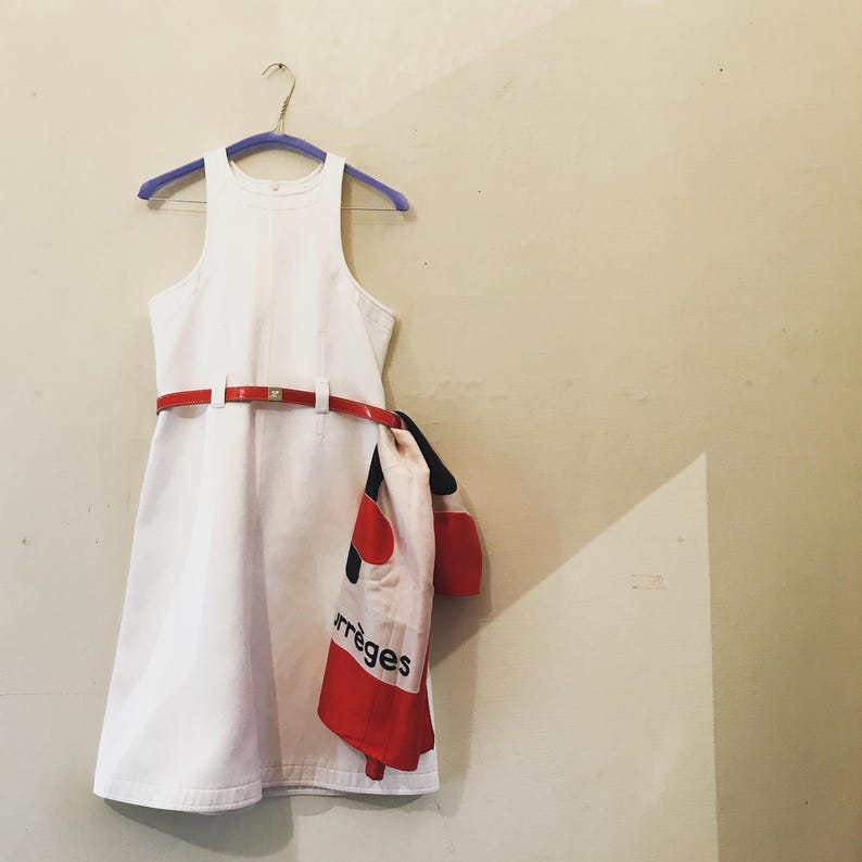 Puede incluir: Un vestido blanco sin mangas con cintur&oacute;n rojo y bufanda a juego. La bufanda tiene detalles en rojo y negro y la palabra "rr&egrave;ges" impresa. El vestido est&aacute; colgado de una percha morada.