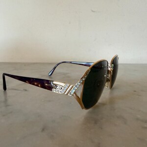 May include: Gold-tone sunglasses featuring dark tinted lenses and a tortoiseshell pattern on the temples. The frame front and hinges are embellished with clear crystals, and the metal temple piece displays the CD logo in a silver-tone finish.