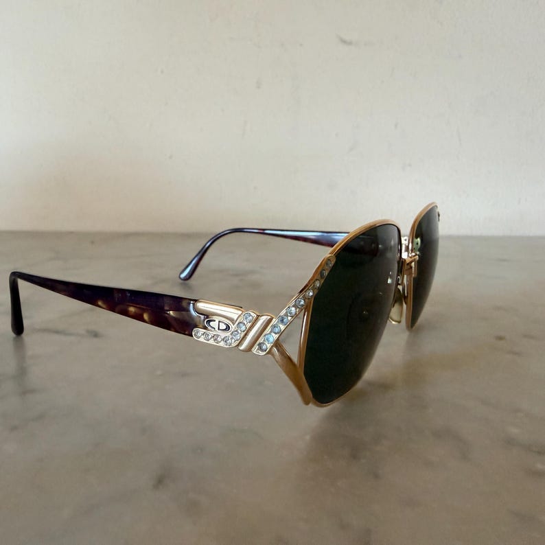 May include: Gold-tone sunglasses featuring dark tinted lenses and a tortoiseshell pattern on the temples. The frame front and hinges are embellished with clear crystals, and the metal temple piece displays the CD logo in a silver-tone finish.