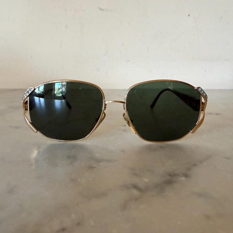 May include: Gold-tone metal sunglasses featuring dark green lenses and a thin wire frame. The outer edges of the frames are accented with a row of small, clear, sparkling crystals embedded into the metal, providing a decorative detail.