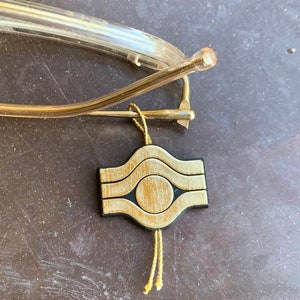 May include: A gold-colored metal eyeglass holder with a wooden charm. The charm is a square with rounded corners and features a circle in the center. The charm is attached to a gold-colored cord with tassels.