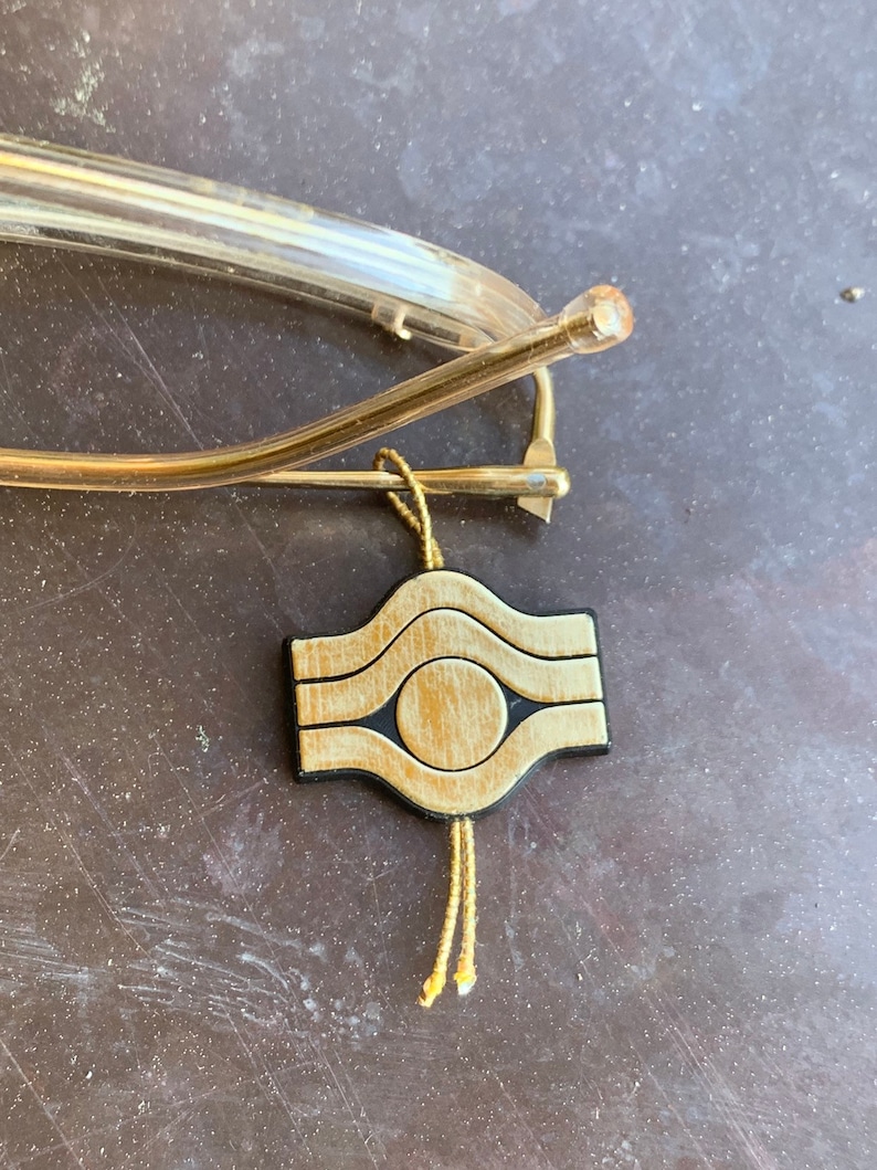 May include: A gold-colored metal eyeglass holder with a wooden charm. The charm is a square with rounded corners and features a circle in the center. The charm is attached to a gold-colored cord with tassels.