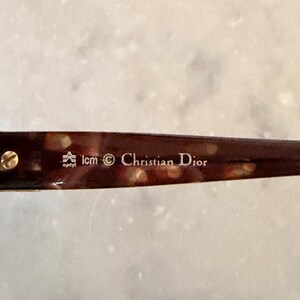 May include: A brown tortoiseshell patterned eyeglass temple arm featuring a gold-toned metal screw and white printed text that reads optyl, lcm, a copyright symbol, and Christian Dior. The material has a glossy finish with visible light reflections.
