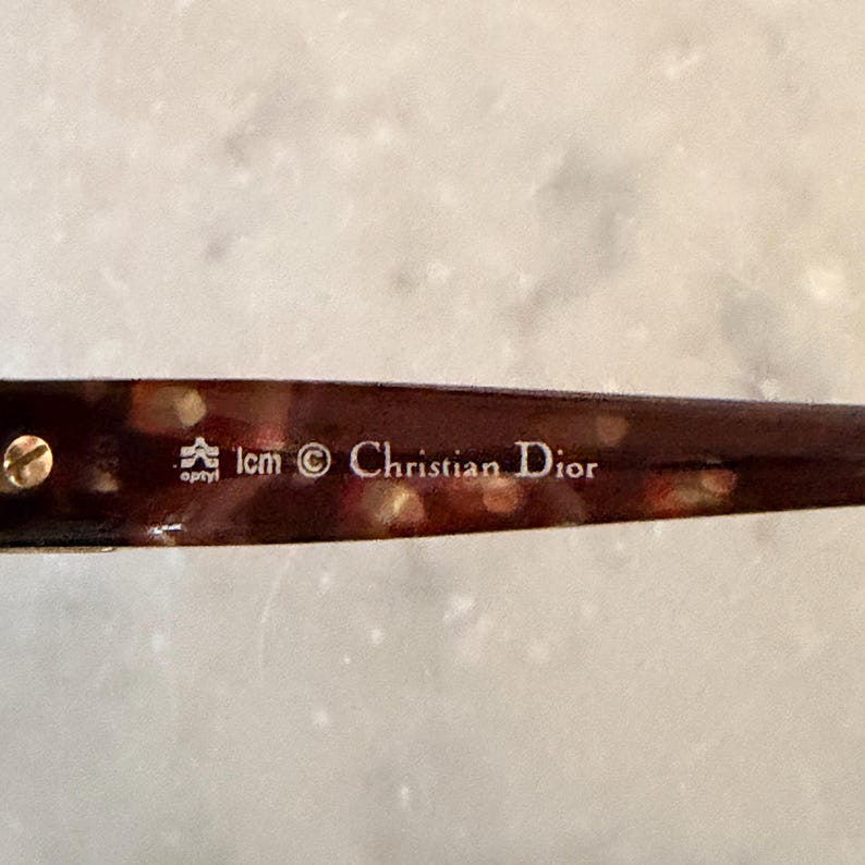 May include: A brown tortoiseshell patterned eyeglass temple arm featuring a gold-toned metal screw and white printed text that reads optyl, lcm, a copyright symbol, and Christian Dior. The material has a glossy finish with visible light reflections.