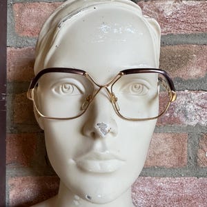 May include: Eyeglasses featuring a gold-tone metal frame with dark brown accents along the upper brow line. The lenses are large and rectangular with rounded corners, connected by a thin gold bridge. The frame includes adjustable nose pads.