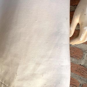 Puede incluir: Primer plano de una tela de color claro, posiblemente una prenda, con una superficie ligeramente texturizada. La tela parece ser de color liso, con sutiles variaciones de tono. El dobladillo inferior es visible, y una mano de maniqu&iacute; est&aacute; en el lado derecho.