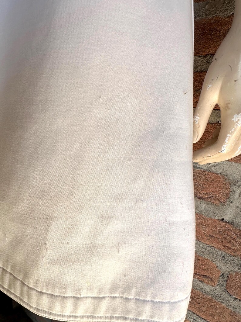 Puede incluir: Primer plano de una tela de color claro, posiblemente una prenda, con una superficie ligeramente texturizada. La tela parece ser de color liso, con sutiles variaciones de tono. El dobladillo inferior es visible, y una mano de maniqu&iacute; est&aacute; en el lado derecho.
