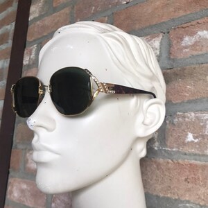 May include: Gold-tone sunglasses with dark lenses and a decorative frame featuring small clear crystals along the hinges. The temples are a dark brown tortoiseshell pattern, and the metal bridge connects the two rounded lenses in a classic design.