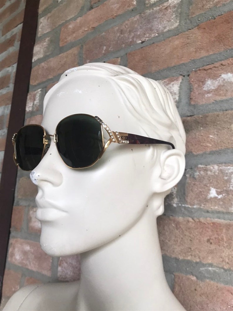 May include: Gold-tone sunglasses with dark lenses and a decorative frame featuring small clear crystals along the hinges. The temples are a dark brown tortoiseshell pattern, and the metal bridge connects the two rounded lenses in a classic design.