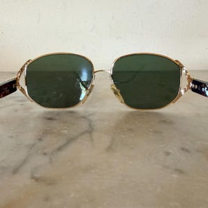 May include: Gold-tone metal sunglasses featuring dark green lenses with a rounded, slightly hexagonal shape. The frame includes intricate, textured gold-tone metal hinges that connect to dark, mottled brown tortoiseshell-patterned temple arms.