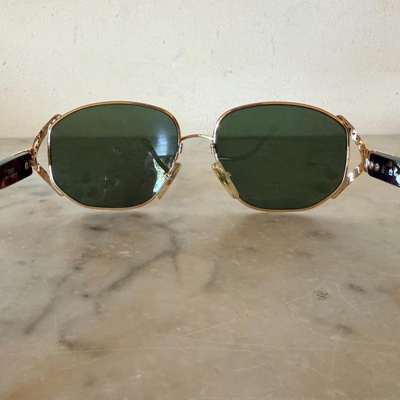 May include: Gold-tone metal sunglasses featuring dark green lenses with a rounded, slightly hexagonal shape. The frame includes intricate, textured gold-tone metal hinges that connect to dark, mottled brown tortoiseshell-patterned temple arms.