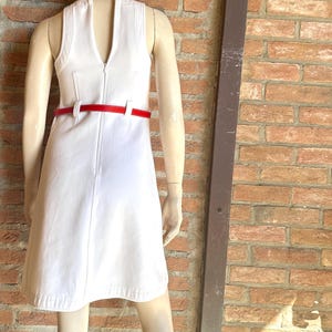 Puede incluir: Un vestido blanco sin mangas con cintur&oacute;n rojo se exhibe en un maniqu&iacute;. El vestido tiene un escote en V y una cremallera en la parte delantera. El cintur&oacute;n rojo ci&ntilde;e la cintura. El maniqu&iacute; est&aacute; frente a una pared de ladrillos.