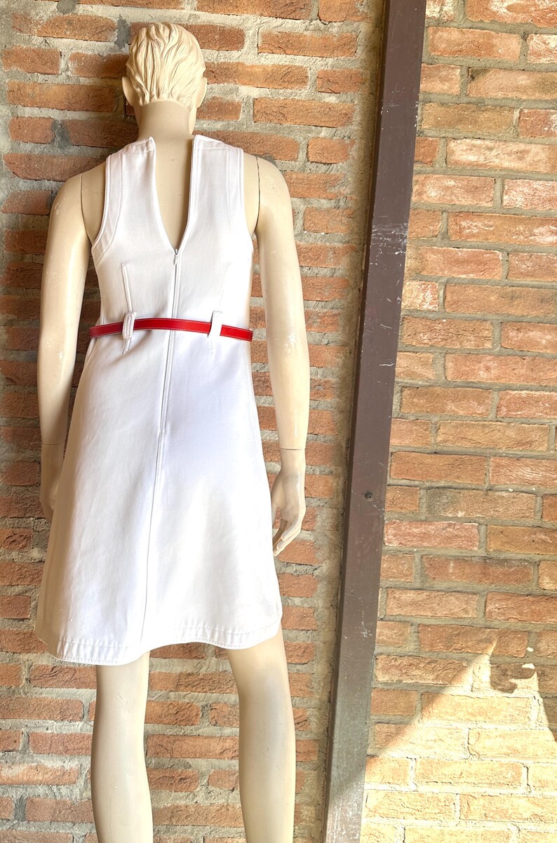 Puede incluir: Un vestido blanco sin mangas con cintur&oacute;n rojo se exhibe en un maniqu&iacute;. El vestido tiene un escote en V y una cremallera en la parte delantera. El cintur&oacute;n rojo ci&ntilde;e la cintura. El maniqu&iacute; est&aacute; frente a una pared de ladrillos.