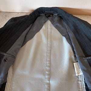 May include: A dark gray pinstripe blazer with a light gray interior lining. The blazer is open, revealing the inside construction and pockets. The lining is a solid light gray color, and the exterior fabric has a subtle pinstripe pattern.