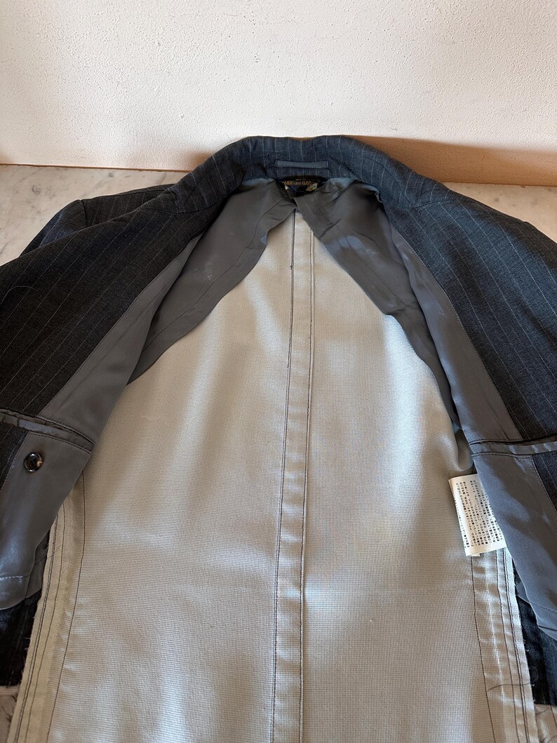 May include: A dark gray pinstripe blazer with a light gray interior lining. The blazer is open, revealing the inside construction and pockets. The lining is a solid light gray color, and the exterior fabric has a subtle pinstripe pattern.