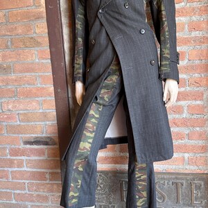 May include: A gray pinstripe suit with camouflage accents. The double-breasted coat and matching pants feature a military-style camouflage pattern on the sleeves and pant legs. The suit is displayed on a mannequin.