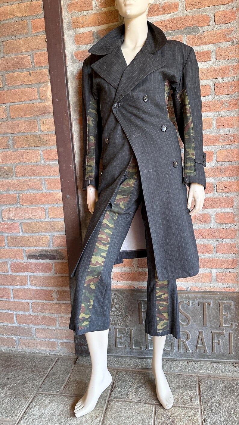 May include: A gray pinstripe suit with camouflage accents. The double-breasted coat and matching pants feature a military-style camouflage pattern on the sleeves and pant legs. The suit is displayed on a mannequin.