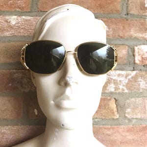 May include: Gold-tone sunglasses with dark tinted lenses and a thin metal frame. The temples feature small, clear embedded crystals on the outer edges near the hinges. The glasses are displayed on a white mannequin head against a brick wall.