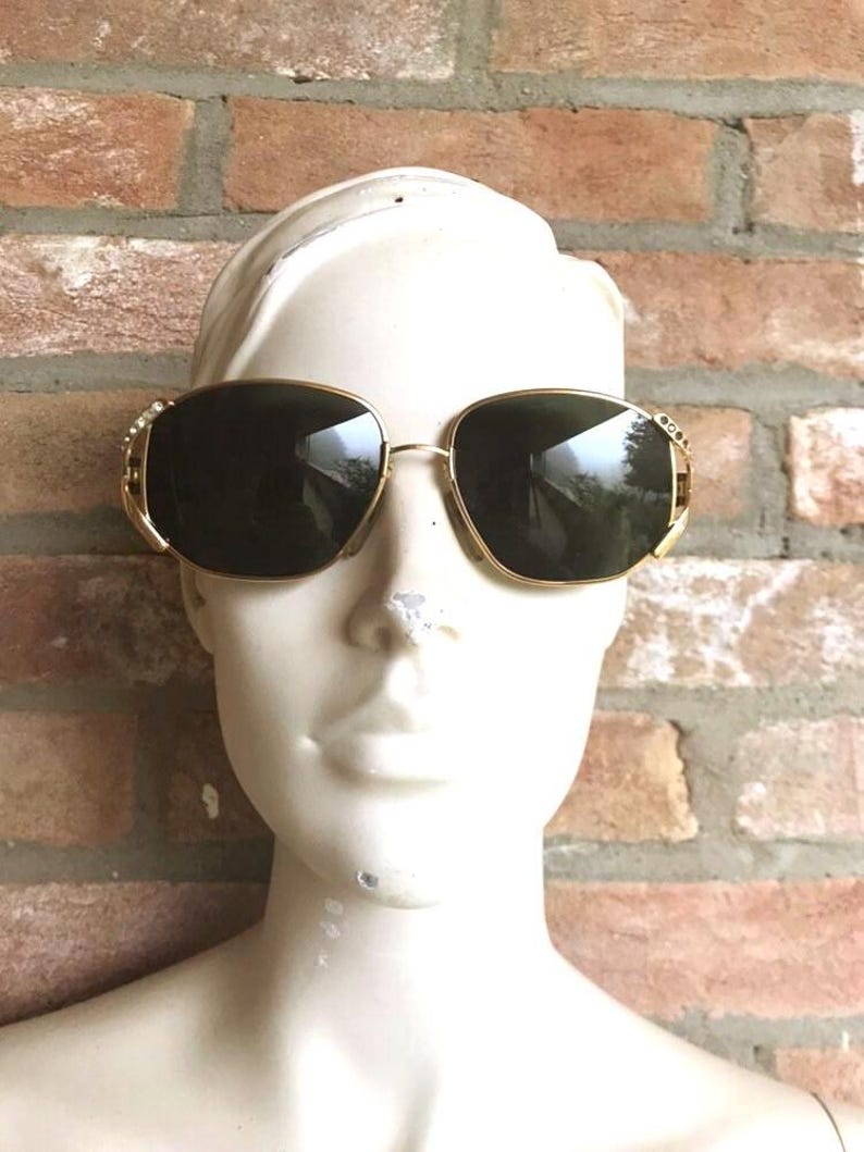 May include: Gold-tone sunglasses with dark tinted lenses and a thin metal frame. The temples feature small, clear embedded crystals on the outer edges near the hinges. The glasses are displayed on a white mannequin head against a brick wall.