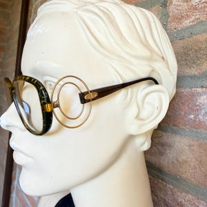 May include: Eyeglasses with dark green frames featuring gold-colored flecks and clear lenses. The temples are dark brown and connect to the frame front via two concentric gold-toned circular metal rings on each side, with a small logo detail.
