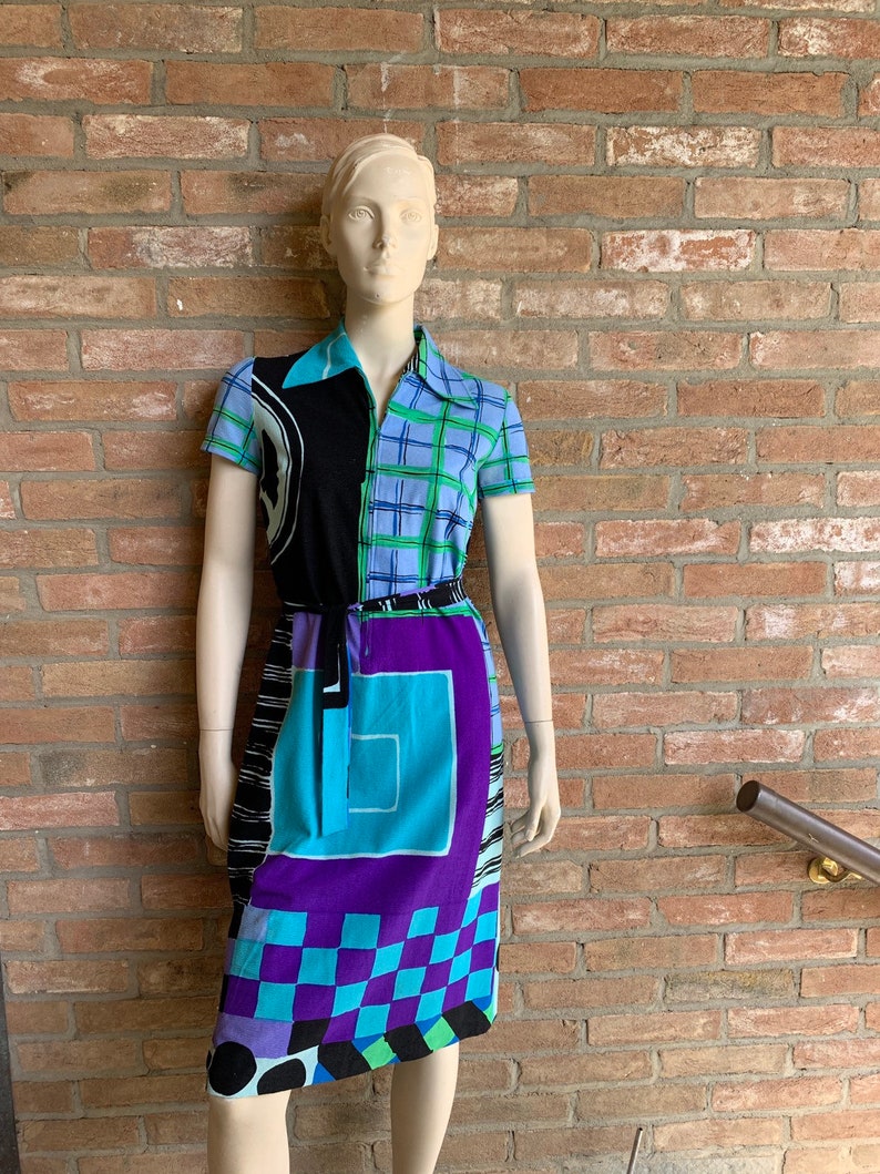 Puede incluir: Un vestido estampado en azul, negro, verde y morado con cuello y mangas cortas. El vestido tiene un dise&ntilde;o geom&eacute;trico y un cintur&oacute;n.