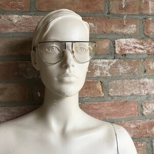 May include: Gold-rimmed eyeglasses with a dark blue top bar and clear lenses. The glasses are displayed on a mannequin against a brick wall background. The glasses have a retro, vintage style.