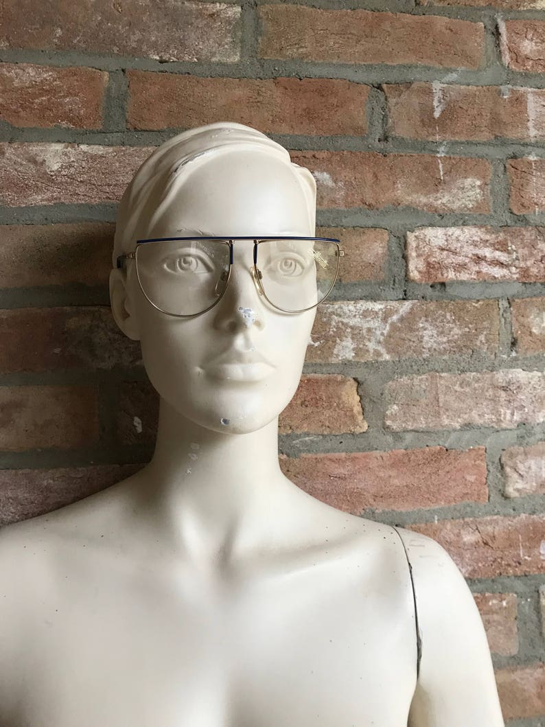 May include: Gold-rimmed eyeglasses with a dark blue top bar and clear lenses. The glasses are displayed on a mannequin against a brick wall background. The glasses have a retro, vintage style.