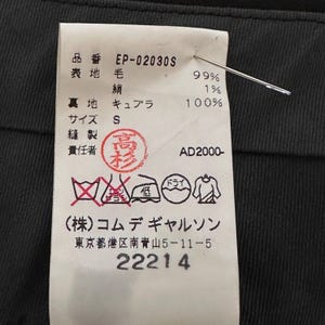 Peut inclure: Gros plan sur une &eacute;tiquette de v&ecirc;tement avec du texte japonais et le num&eacute;ro "22214". L'&eacute;tiquette indique la composition du tissu : 99% laine, 1% soie et 100% cupro. Une aiguille perce l'&eacute;tiquette, fix&eacute;e &agrave; un tissu sombre.