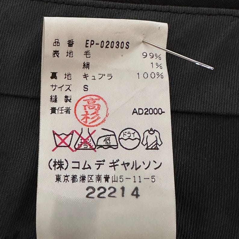 Peut inclure: Gros plan sur une &eacute;tiquette de v&ecirc;tement avec du texte japonais et le num&eacute;ro "22214". L'&eacute;tiquette indique la composition du tissu : 99% laine, 1% soie et 100% cupro. Une aiguille perce l'&eacute;tiquette, fix&eacute;e &agrave; un tissu sombre.