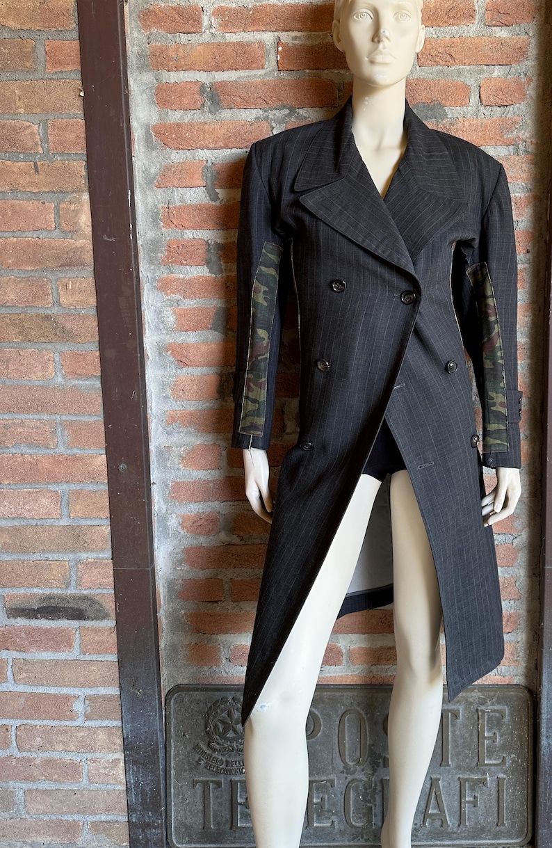 May include: A dark gray pinstripe double-breasted coat with camouflage patterned sleeves. The coat is displayed on a mannequin against a brick wall background. The coat has a long, asymmetrical design.