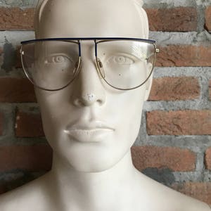 May include: Gold-rimmed eyeglasses with a blue top bar and clear lenses. The glasses are displayed on a white mannequin head against a brick wall background. The brand name "Silhouette" is visible on the lens.