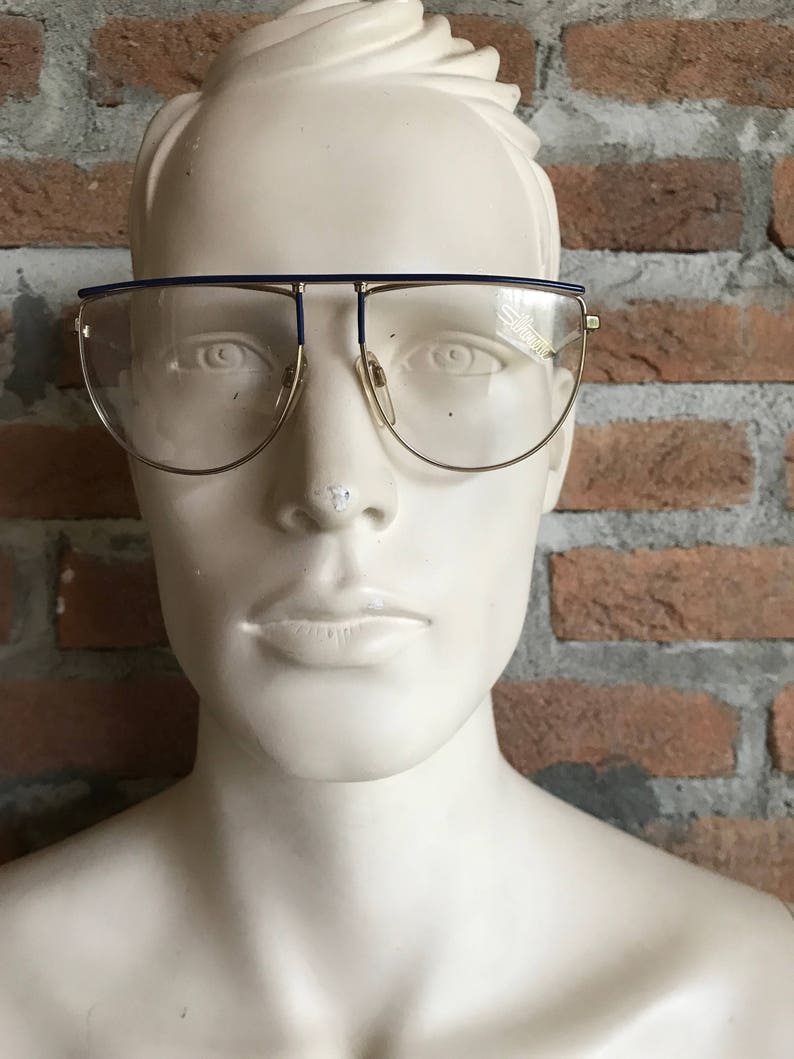 May include: Gold-rimmed eyeglasses with a blue top bar and clear lenses. The glasses are displayed on a white mannequin head against a brick wall background. The brand name "Silhouette" is visible on the lens.