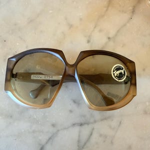 Peut inclure: Lunettes de soleil surdimensionnées avec une monture dégradée marron et des verres teintés. La branche gauche porte une étiquette argentée indiquant MOD. 2754, et le verre droit présente un autocollant circulaire avec le texte Garantito Temperal Anti Choc.