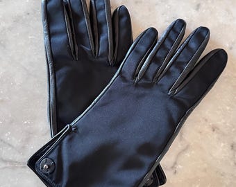 May include: A pair of black gloves with a satin finish. The gloves have a leather trim and a button closure at the wrist. The gloves are lying on a marble surface.