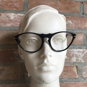 May include: Black eyeglasses with a rounded frame shape and a high bridge. The frames feature thin black rims and small gold-tone accents at the outer corners where the temples connect to the front of the glasses, displayed on a mannequin head.