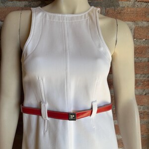 Puede incluir: Un vestido blanco sin mangas con cintur&oacute;n rojo. El vestido tiene costuras verticales y cuello alto. El cintur&oacute;n rojo tiene hebilla plateada con logo. El vestido se exhibe en un maniqu&iacute;.