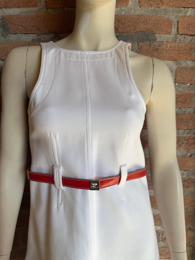 Puede incluir: Un vestido blanco sin mangas con cintur&oacute;n rojo. El vestido tiene costuras verticales y cuello alto. El cintur&oacute;n rojo tiene hebilla plateada con logo. El vestido se exhibe en un maniqu&iacute;.