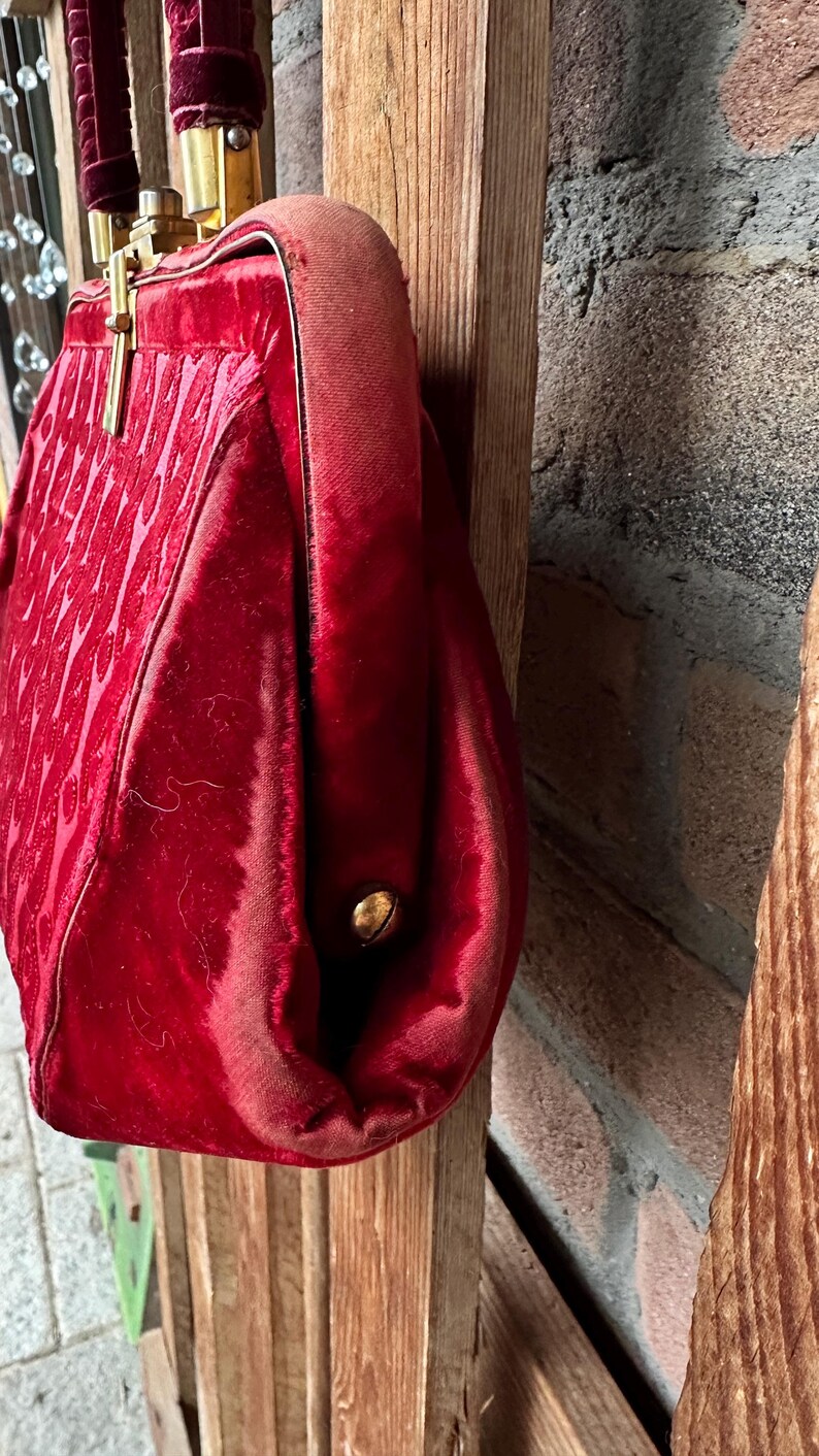 Pu&ograve; includere: Una borsa a mano in velluto rosso intenso con un motivo testurizzato sul davanti, una chiusura a telaio in metallo dorato e un manico in velluto abbinato. Il lato della borsa &egrave; fissato con un piccolo rivetto in metallo dorato e tessuto.