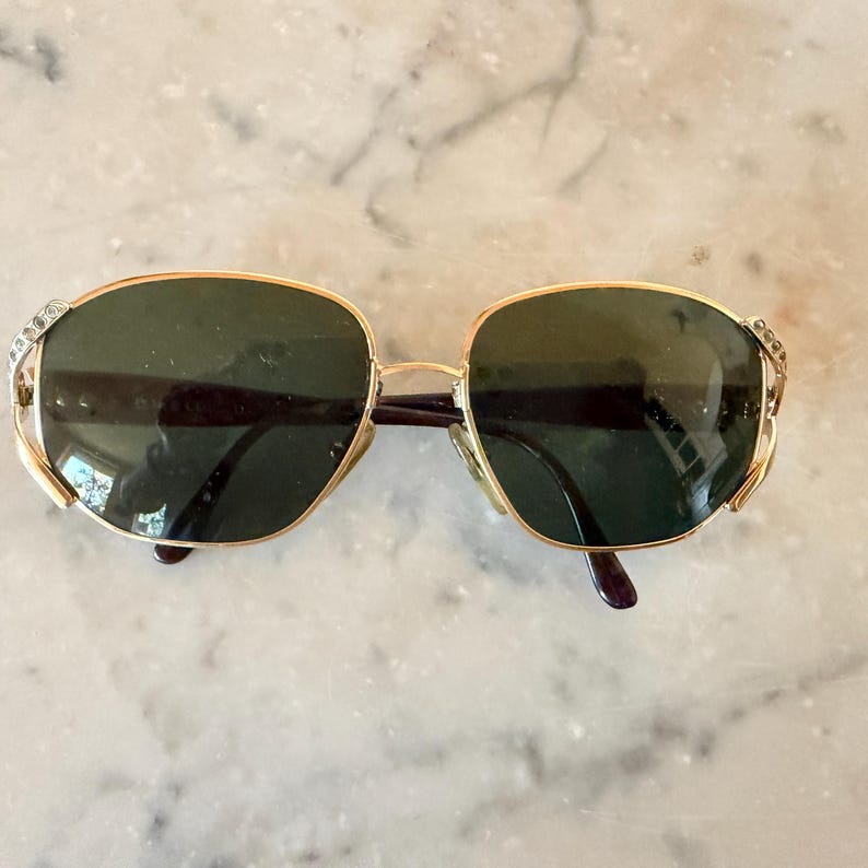 May include: Gold-tone sunglasses featuring oval-shaped dark green lenses and a thin metal frame. The hinges are accented with small, clear embedded crystals on each side, and the temples are finished with dark brown tips for a secure fit.