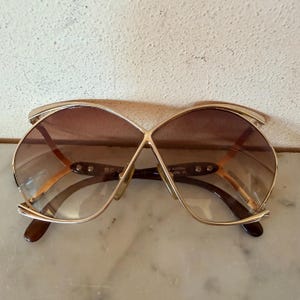 May include: Oversized sunglasses featuring a gold-tone metal frame with a distinctive cross-over bridge design. The lenses are gradient brown, transitioning from darker at the top to lighter at the bottom. The temples are dark brown with visible metal rivets.