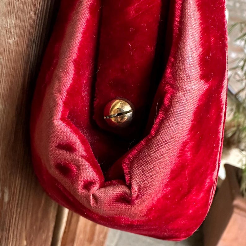 Pu&ograve; includere: Una pochette in velluto rosso intenso caratterizzata da una consistenza morbida e vellutata e da una silhouette arrotondata. La borsa &egrave; chiusa da un singolo bottone a pressione in metallo dorato lucido centrato sulla patta anteriore.