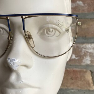 May include: Gold-rimmed eyeglasses with a blue top bar and temples. The lenses are clear, and the brand name "Silhouette" is visible on one lens. The glasses are displayed on a white mannequin head against a brick background.