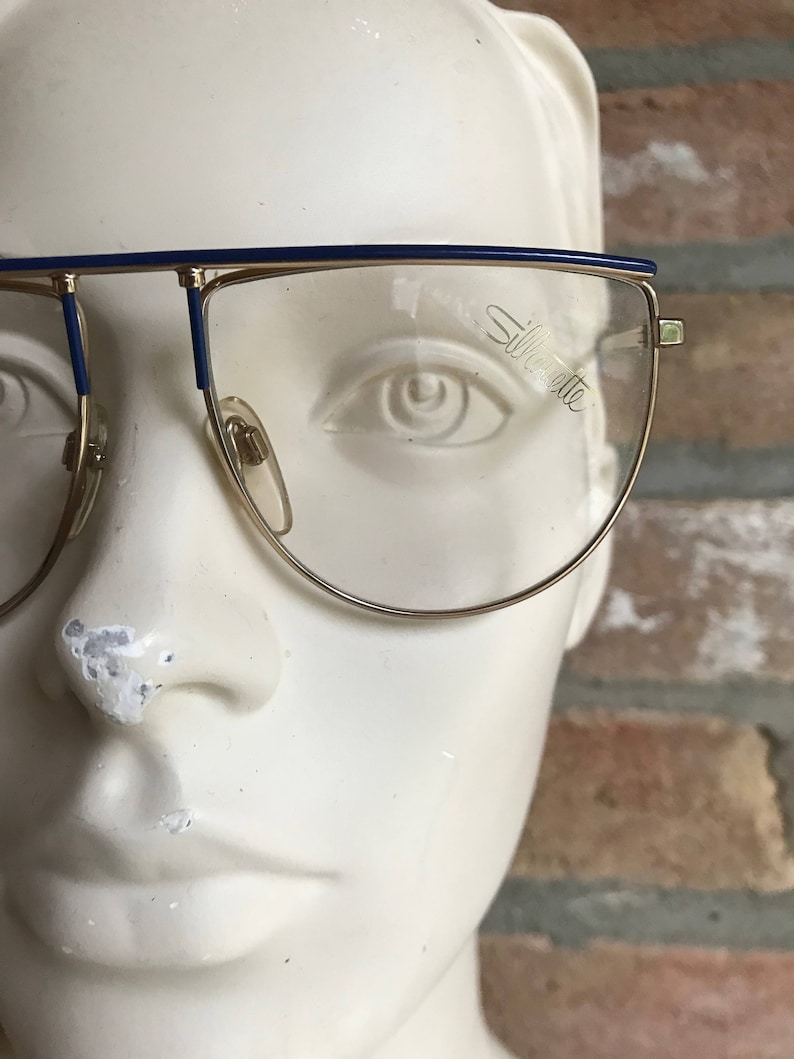 May include: Gold-rimmed eyeglasses with a blue top bar and temples. The lenses are clear, and the brand name "Silhouette" is visible on one lens. The glasses are displayed on a white mannequin head against a brick background.