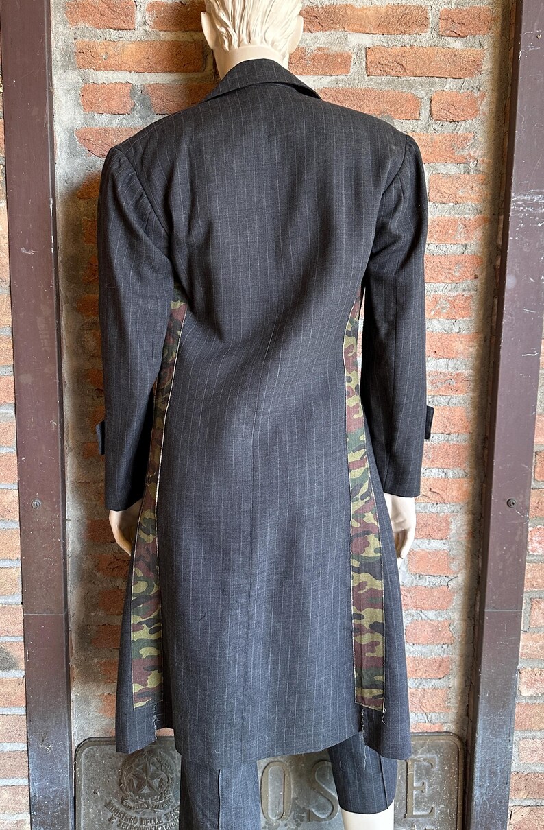 May include: A dark gray pinstripe coat with camouflage side panels. The long coat has a notched lapel, long sleeves, and a tailored fit. The camouflage pattern features shades of green, brown, and tan.