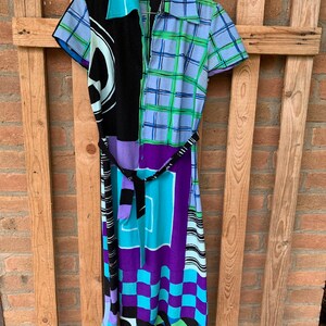 Puede incluir: Un vestido colorido con estampado geom&eacute;trico y cintur&oacute;n negro. El vestido tiene cuello y mangas cortas. El estampado incluye negro, azul, verde, morado y blanco.