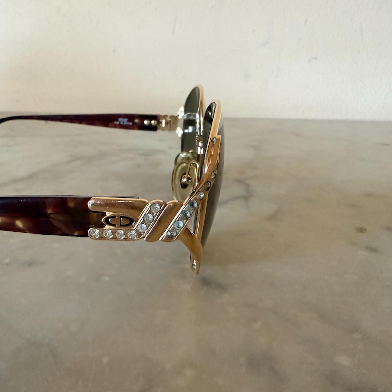 May include: Sunglasses with dark brown tortoiseshell temples and gold-tone metal accents. The hinges feature a CD logo surrounded by clear crystals. The frame front is gold-tone metal with dark lenses, showing a side profile view of the eyewear.