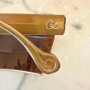 May include: A pair of brown tortoiseshell-patterned eyeglasses featuring a gold-toned cursive logo on the temple arm. The temple arm has a distinct curved, swirl-shaped end piece, and the frame is constructed with a smooth, polished finish.